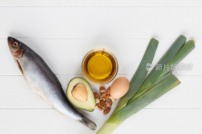 选择含有omega - 3和不饱和脂肪的食物来源。欧米茄3，欧米茄6。含有健康脂肪的产品:亚麻籽油，鳄梨，杏仁，腰果，开心果，鱼，鸡蛋，韭菜。俯视图，白色木板